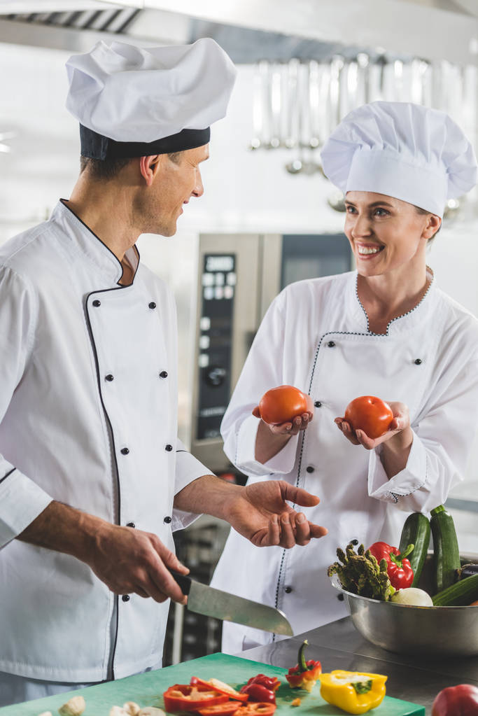 hymyilevä kokki näyttää kypsiä tomaatteja kollegalleen ravintolan keittiössä
 - Valokuva, kuva
