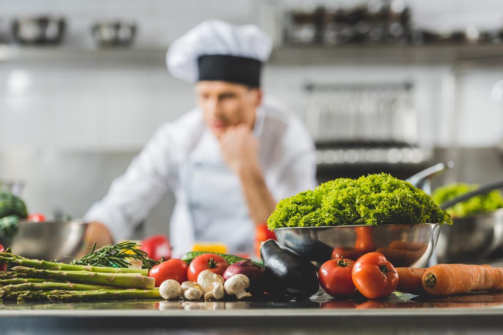 pensive chef at restaurant kitchen with vegetables on foreground - Photo, Image