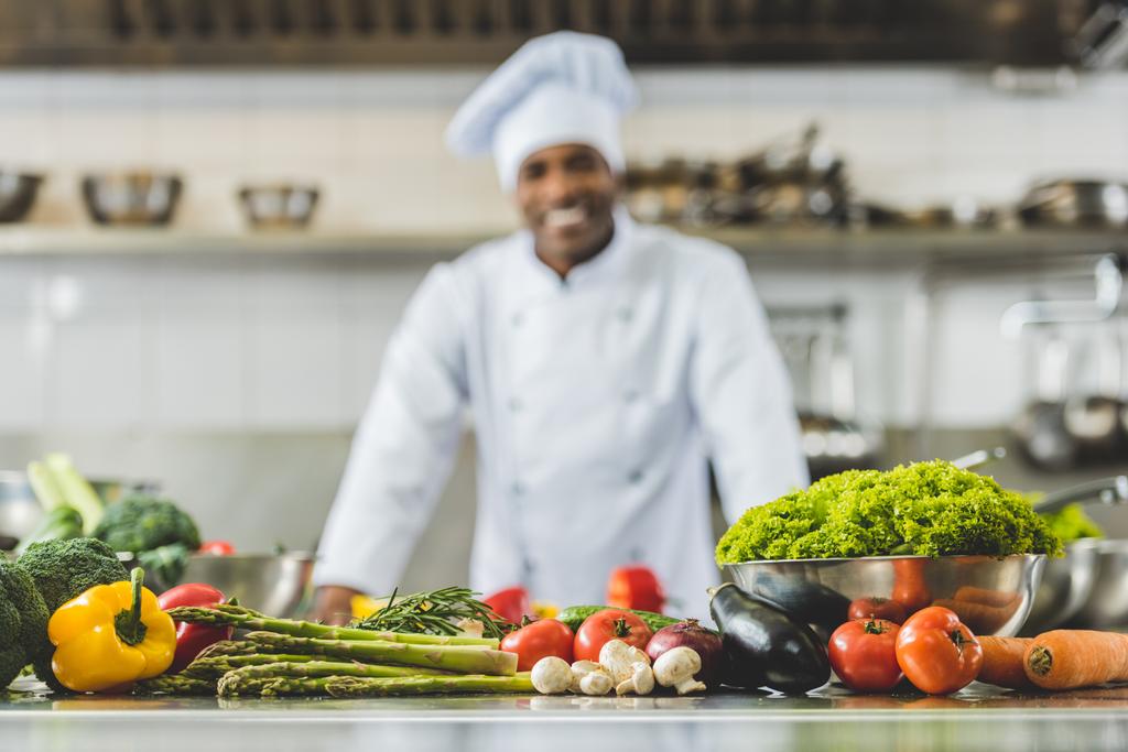 hymyilevä afrikkalainen amerikkalainen kokki ravintolan keittiössä vihannekset etualalla
 - Valokuva, kuva