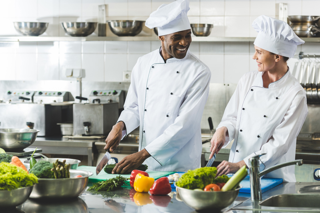 Lächelnde multikulturelle Köche schneiden Gemüse in der Restaurantküche und schauen einander an - Foto, Bild