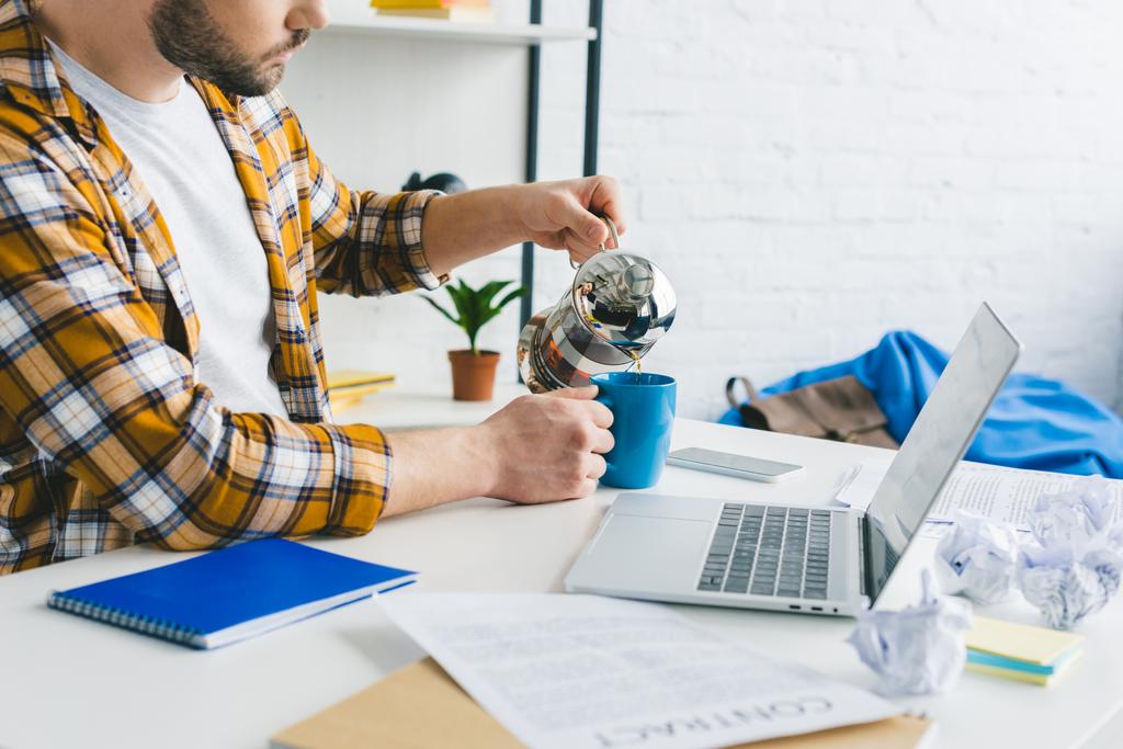 Freelancer derramando café na xícara na mesa com laptop no escritório em casa
 - Foto, Imagem