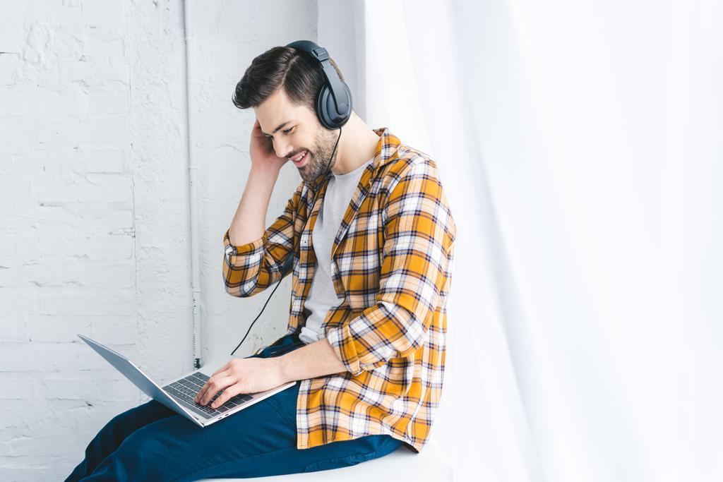 Usmívající se muž v sluchátka pracují na notebooku oknem - Fotografie, Obrázek