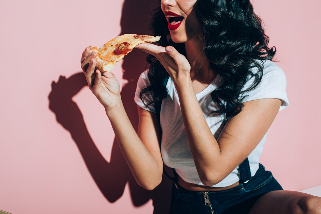 обрезанный снимок молодой женщины, поедающей пиццу на розовом фоне
  - Фото, изображение