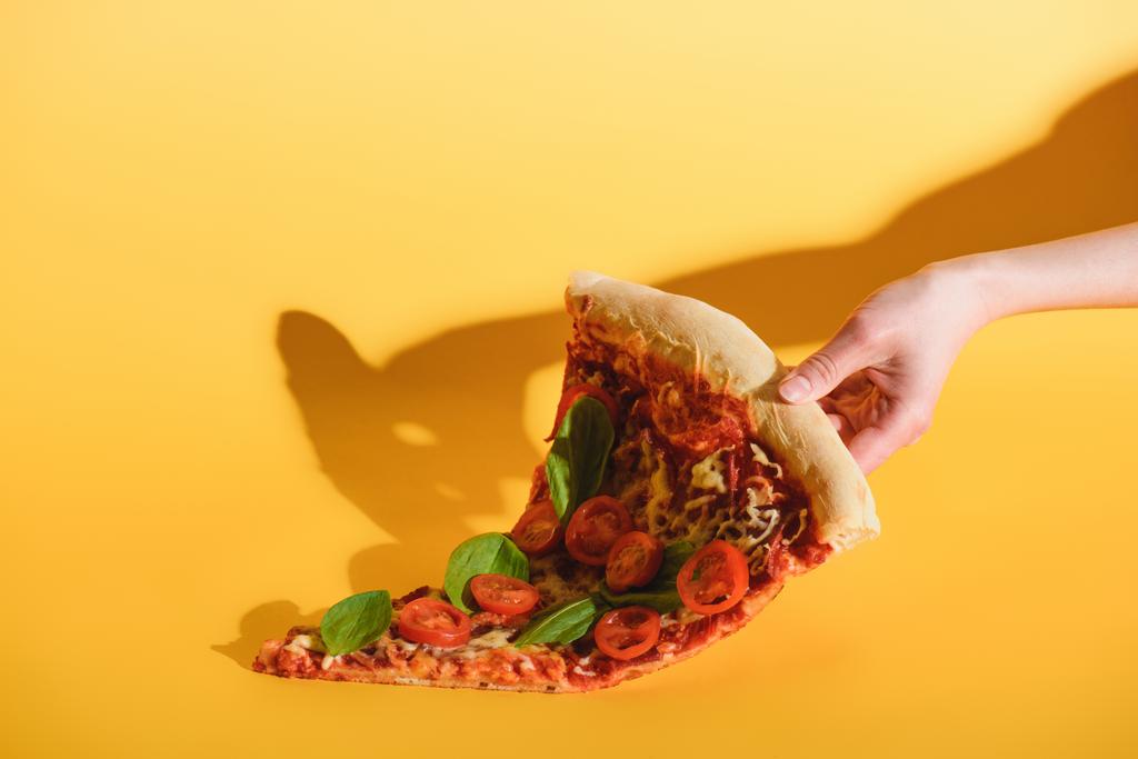 częściowy widok kobieta trzyma kawałek pizzy z pomidorkami w ręku na żółtym tle - Zdjęcie, obraz