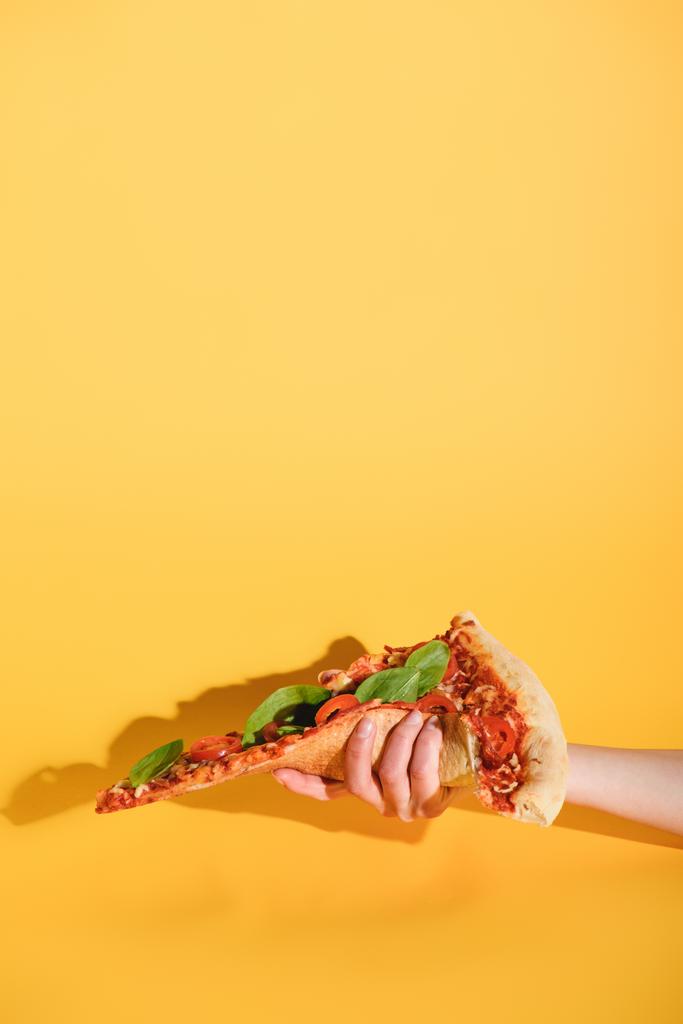 částečný pohled ženy držící kousek pizzy v ruce na žlutém podkladu - Fotografie, Obrázek