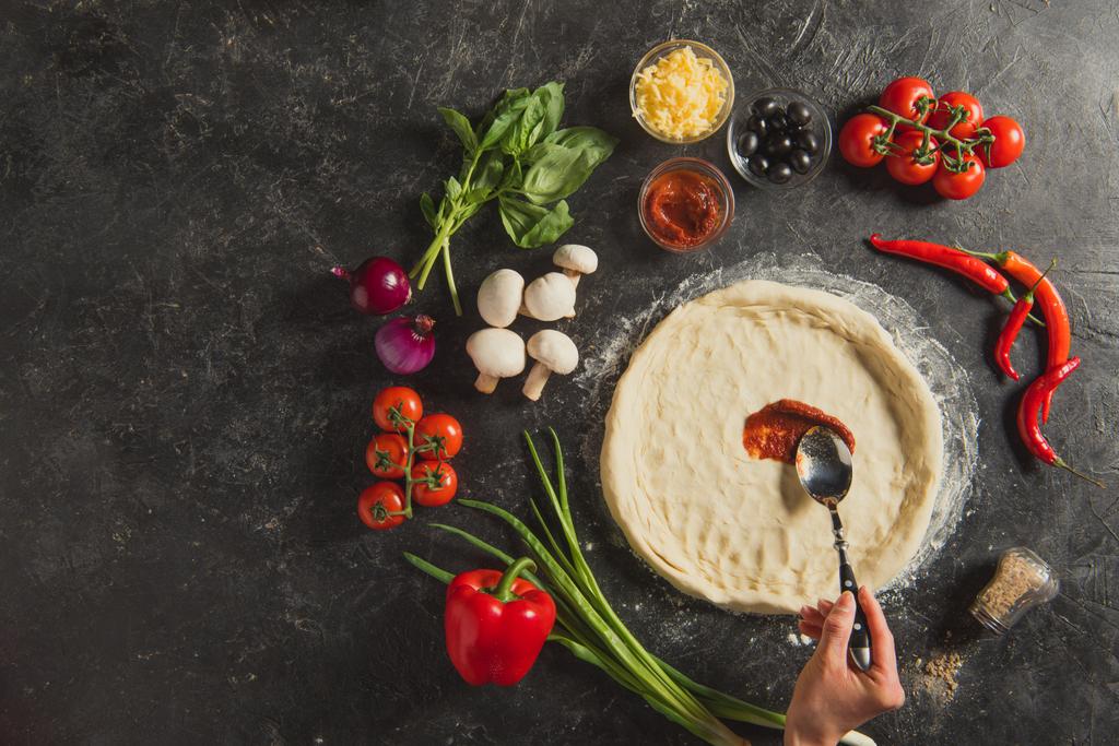 plan recadré de femme mettant de la sauce sur la pâte crue pendant la cuisson de pizza italienne sur table sombre
 - Photo, image