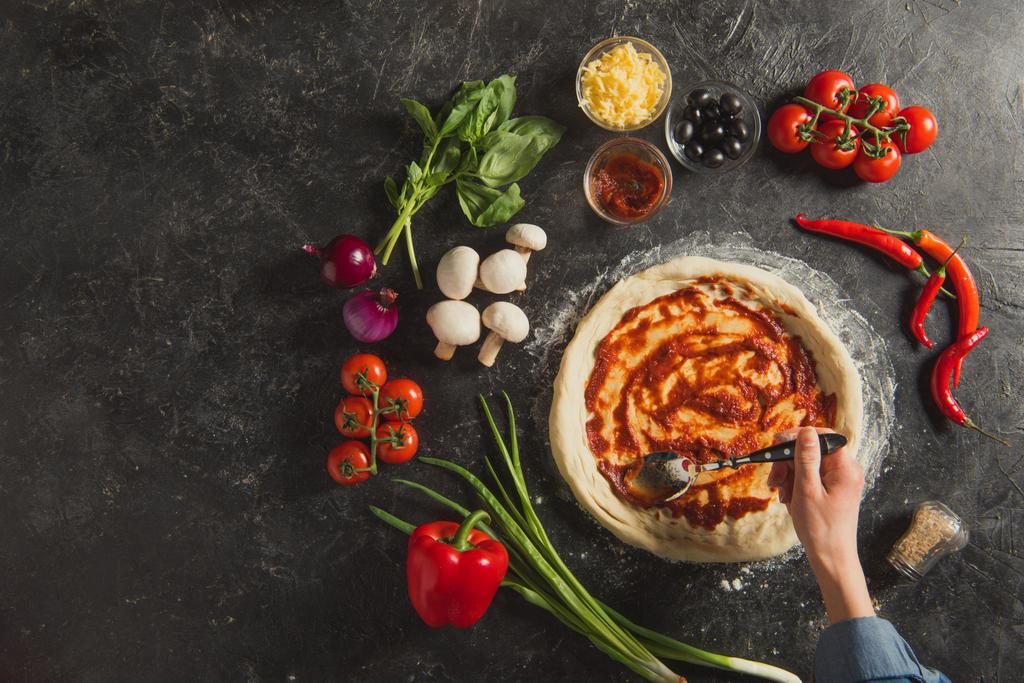 bijgesneden schot van vrouw zetten saus op ruwe deeg tijdens het koken Italiaanse pizza op donkere tafelblad - Foto, afbeelding