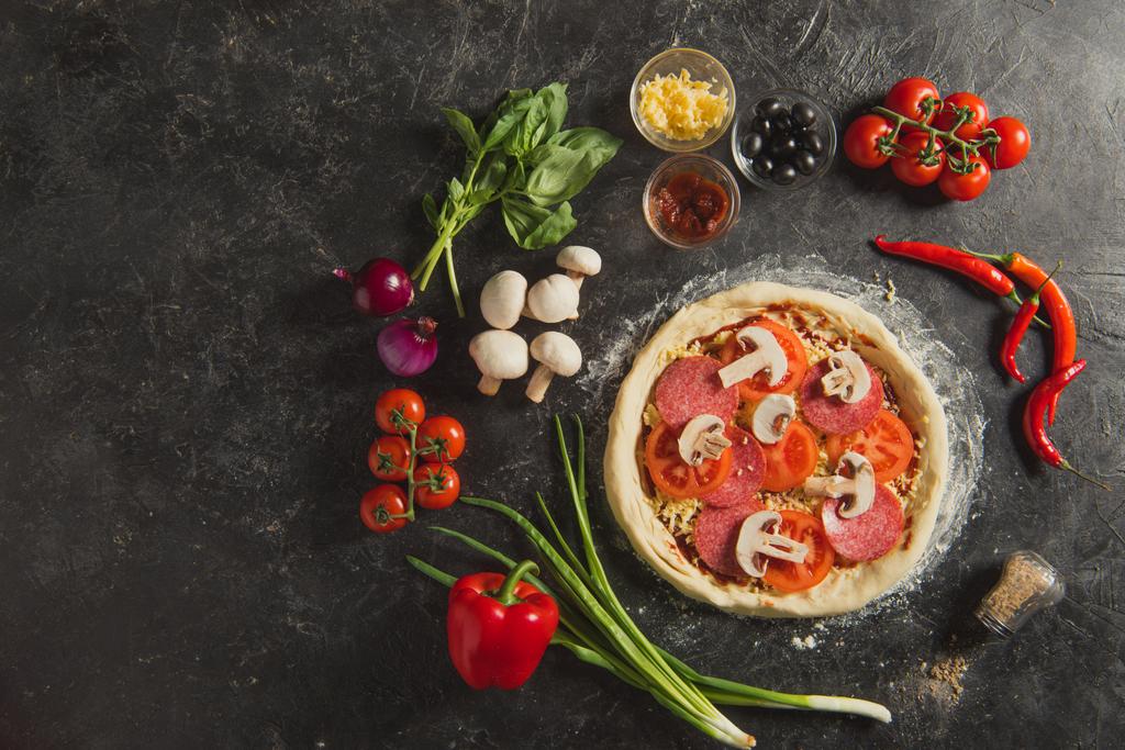 bovenaanzicht van ruwe Italiaanse pizza en verse ingrediënten rond op donkere ondergrond - Foto, afbeelding