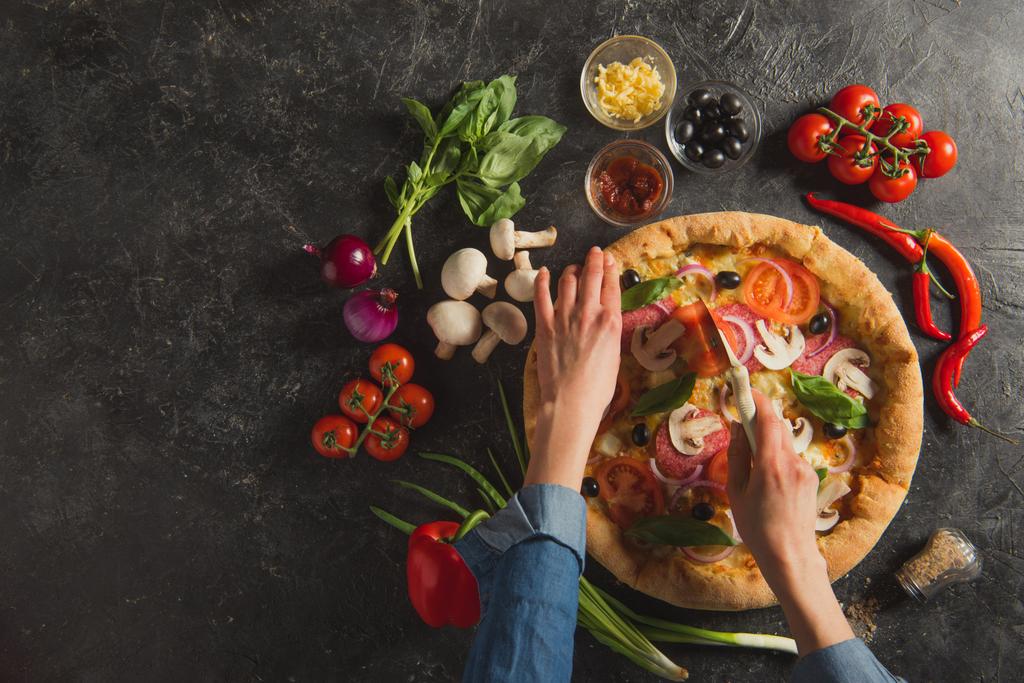 περικοπεί shot γυναίκας κοπή μαγειρεμένα Ιταλική Πίτσα με φρέσκα υλικά σε σκούρο επιτραπέζια - Φωτογραφία, εικόνα