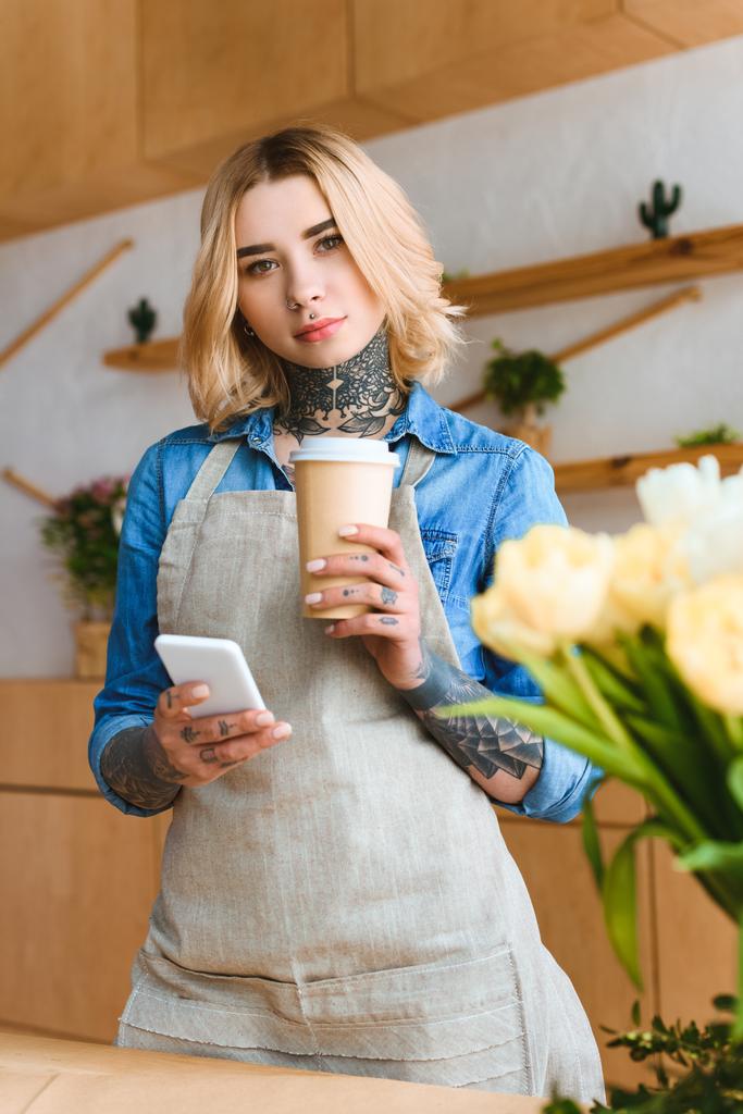 hermosa florista joven sosteniendo café para ir y teléfono inteligente y mirando a la cámara en la tienda de flores
  - Foto, Imagen