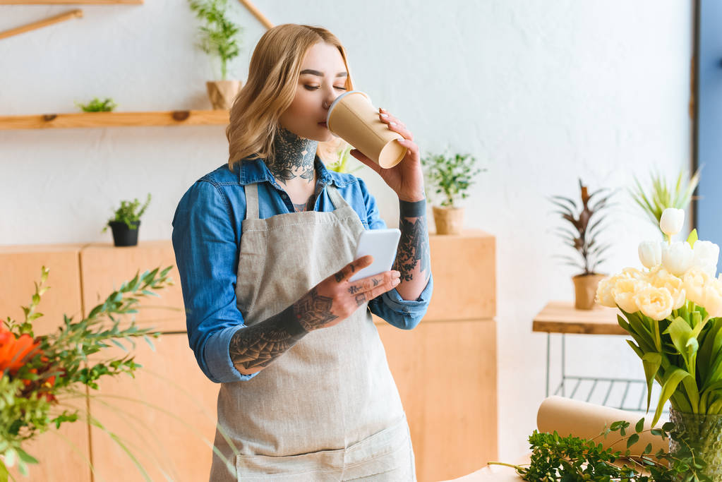 молодой флорист в фартуке пьет кофе из бумажной чашки и пользуется смартфоном в цветочном магазине
 - Фото, изображение