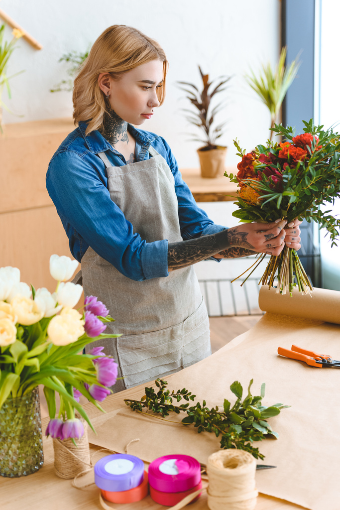 ориентированный молодой флорист в фартуке с красивым букетом в цветочном магазине
 - Фото, изображение