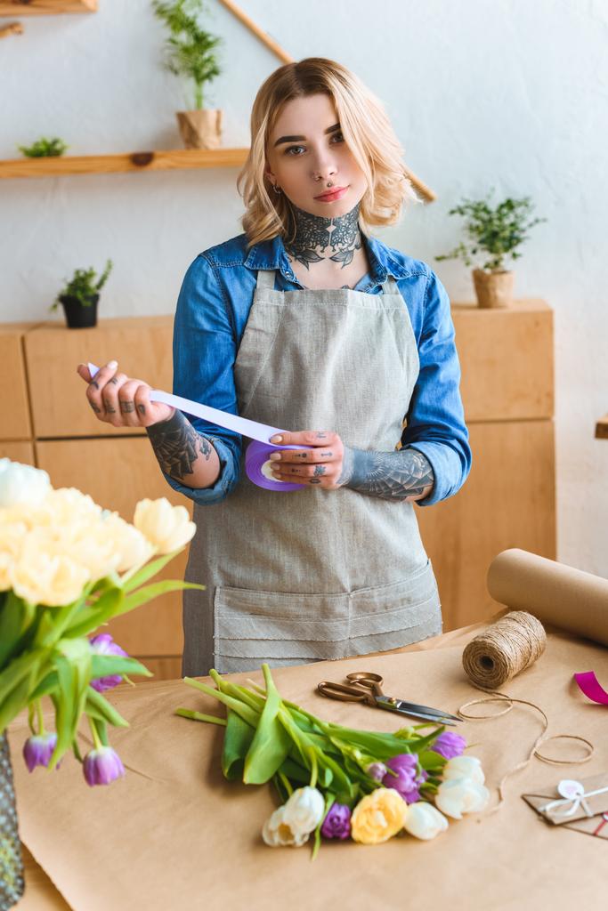 florist holding ribbon and looking at camera while working in flower shop - Photo, Image