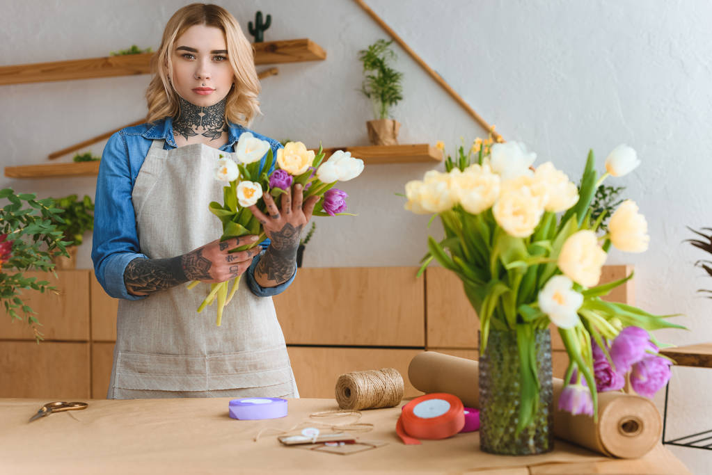 jonge bloemist in schort holding prachtige tulpen en kijken naar de camera in de bloemenwinkel - Foto, afbeelding