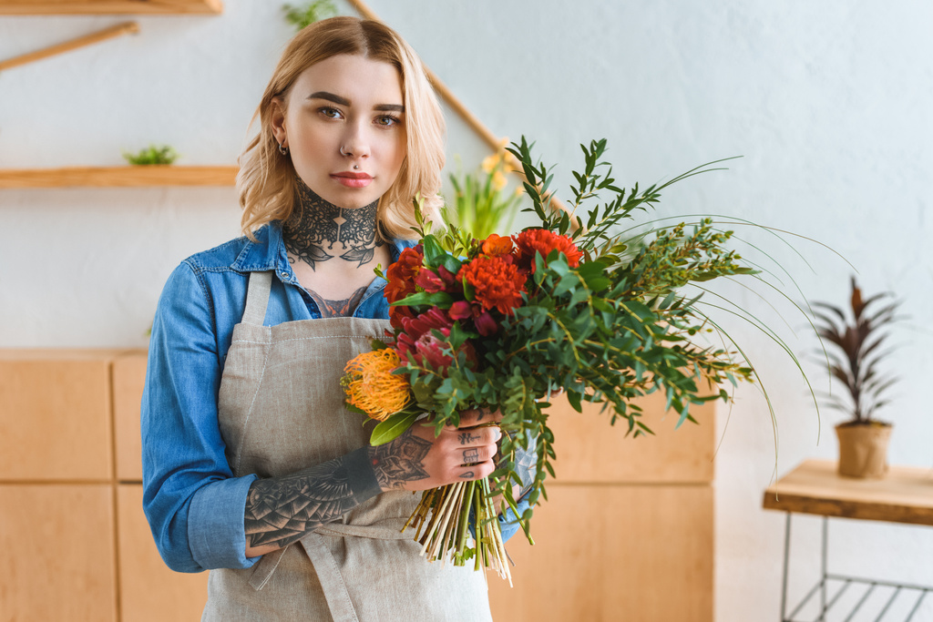 beautiful young florist with tattoos holding flower bouquet and looking at camera - Photo, Image