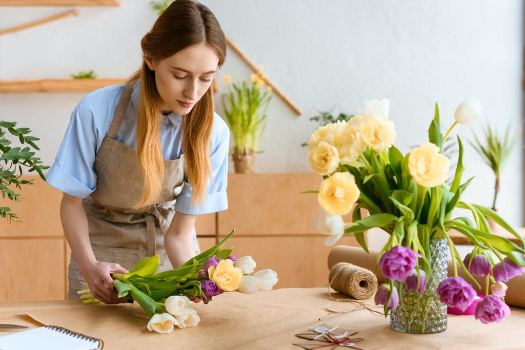 молодой флорист в фартуке устраивает красивые цветы тюльпанов на рабочем месте
 - Фото, изображение