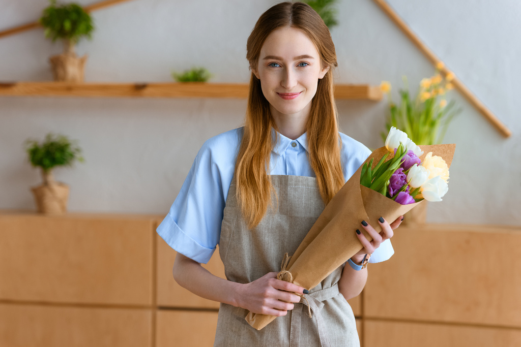 beautiful young florist holding bouquet of tulips and smiling at camera - Photo, Image