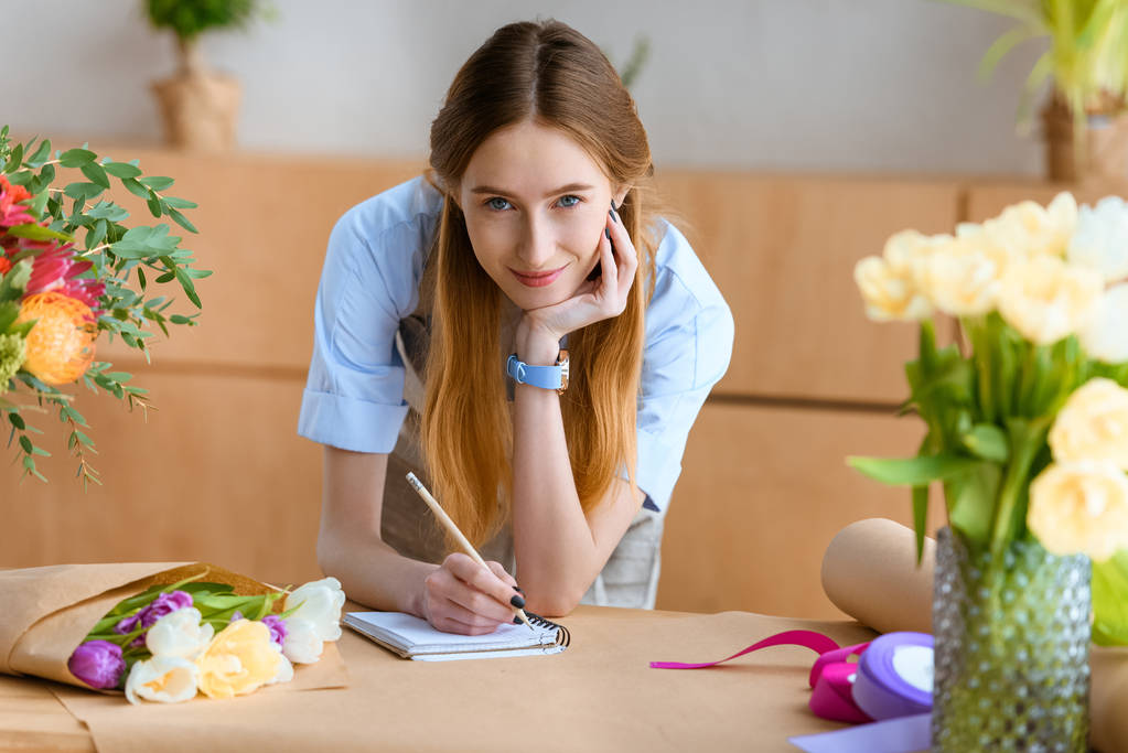 bela jovem florista tomar notas e sorrindo para a câmera no local de trabalho
 - Foto, Imagem