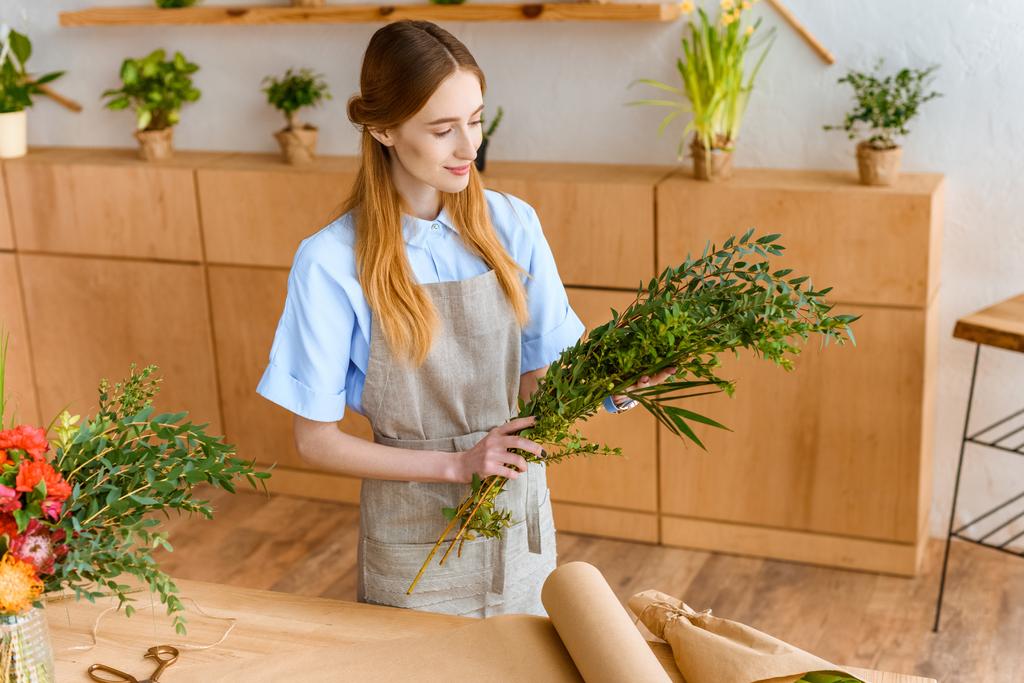 високий кут зору усміхненого молодого флориста в фартусі, що тримає зелені рослини в квітковому магазині
 - Фото, зображення