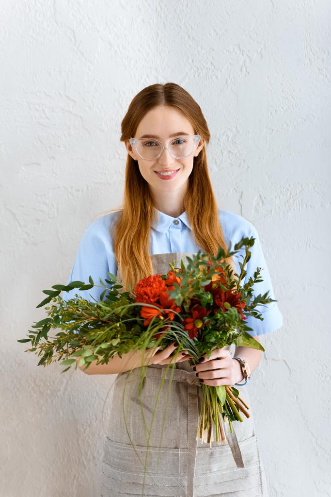 beautiful young florist in eyeglasses hoding flower bouquet and smiling at camera - Photo, Image