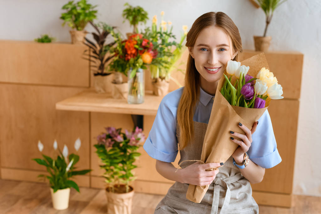 mooie jonge bloemist holding boeket tulpen en glimlachend in de camera in de bloemenwinkel - Foto, afbeelding