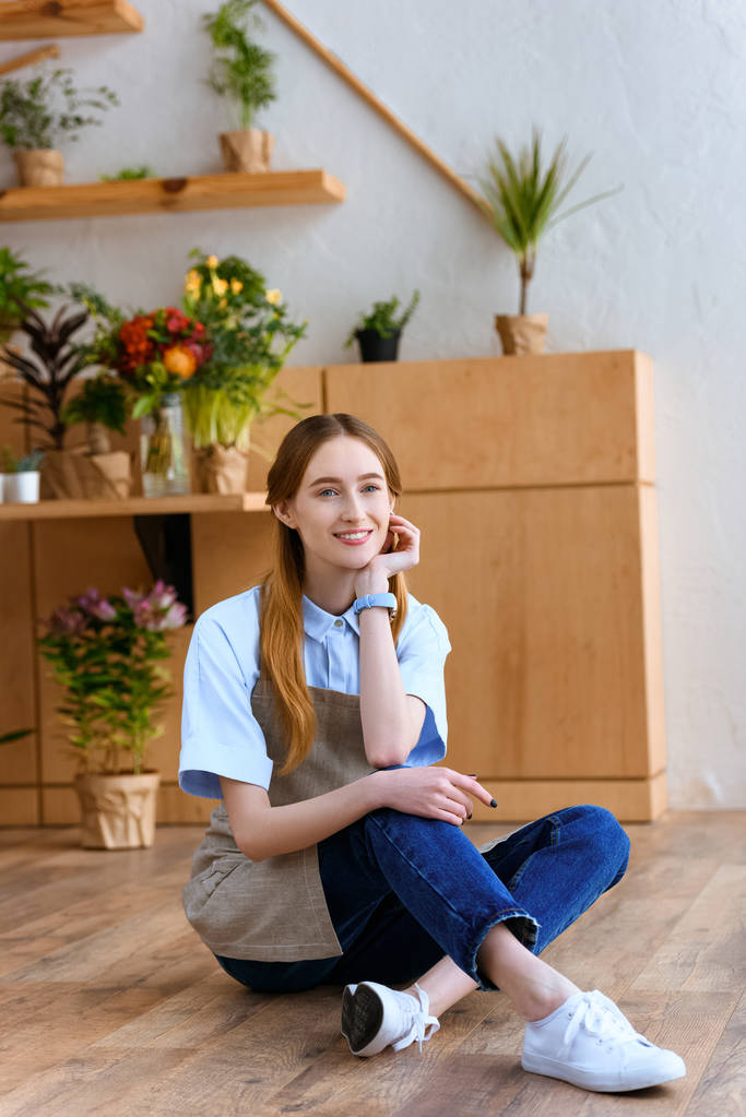beautiful young woman in apron sitting on floor and smiling at camera in flower shop  - Photo, Image