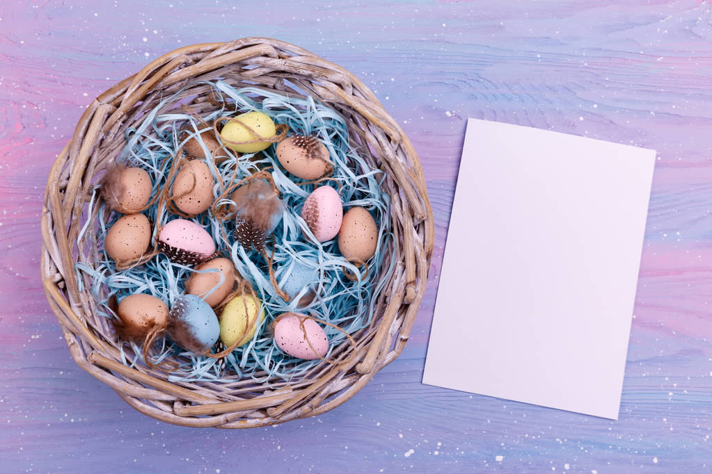 Fondo de Pascua con coloridos huevos de codorniz, plumas en una cesta y un papel vacío cerca de ella, vista superior
.  - Foto, Imagen