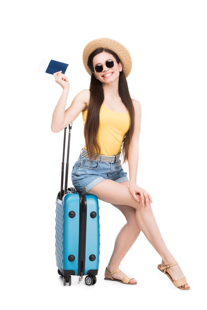 pretty smiling girl with passport, air ticket sitting on travel bag, isolated on white  - Photo, Image