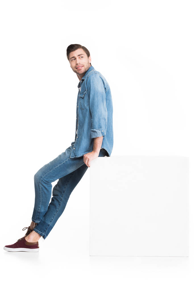 young smiling man in jeans posing at white cube, isolated on white - Photo, Image