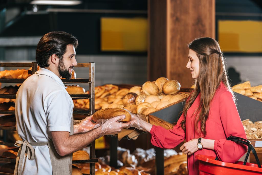 vista lateral do assistente de loja dando pão à mulher com cesta de compras no supermercado
 - Foto, Imagem