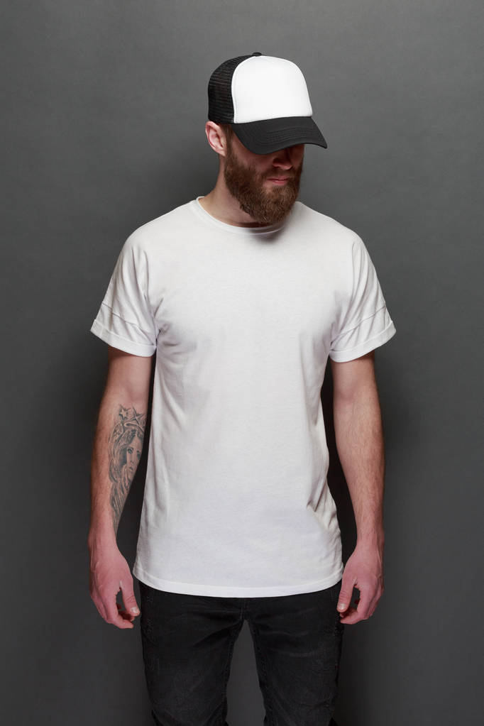 Hipster gutaussehendes männliches Model mit Bart trägt weißes Blank-T-Shirt und Baseballmütze mit Platz für Ihr Logo oder Design im lässigen urbanen Stil - Foto, Bild