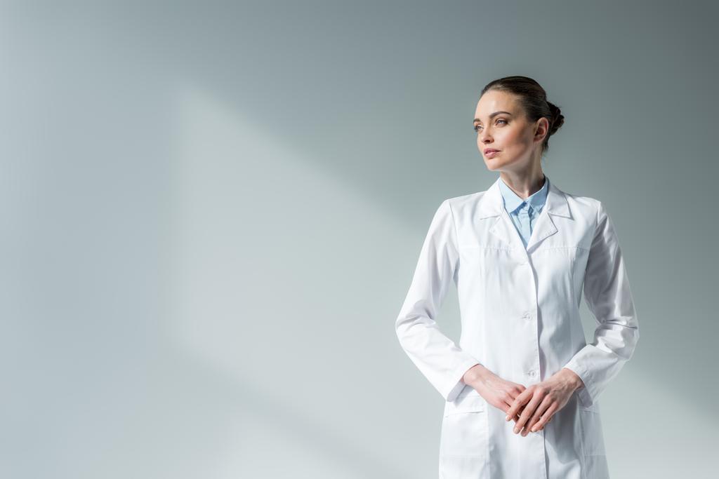 ελκυστική γυναίκα γιατρό σε λευκό παλτό κοιτάζοντας μακριά σε γκρι - Φωτογραφία, εικόνα