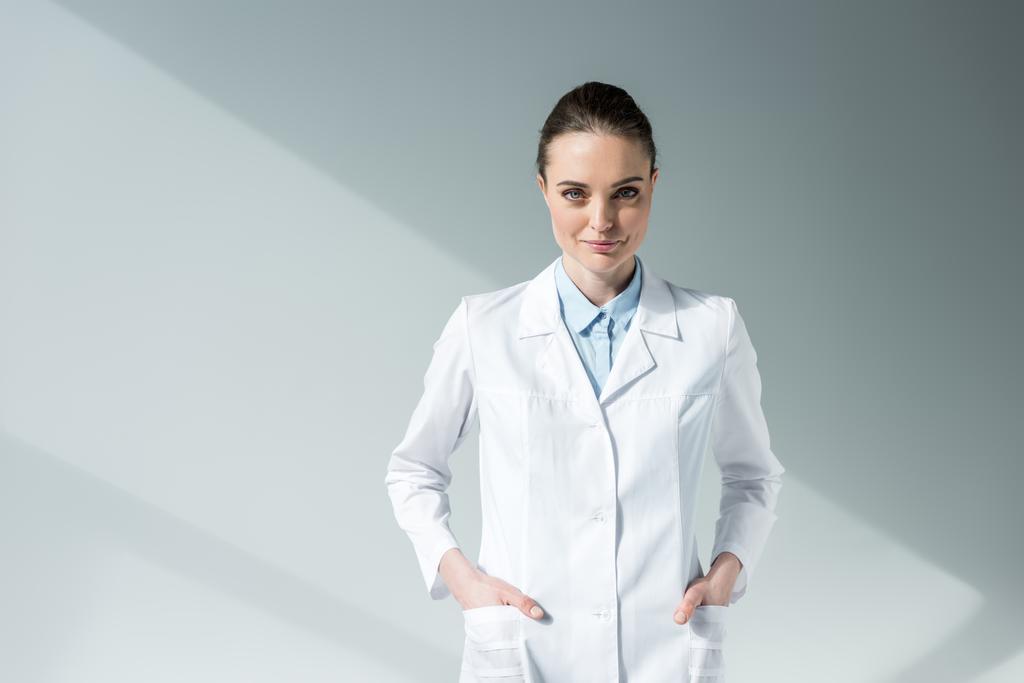 χαμογελαστή γυναίκα γιατρό σε λευκό παλτό κοιτάζοντας την κάμερα στο γκρι - Φωτογραφία, εικόνα