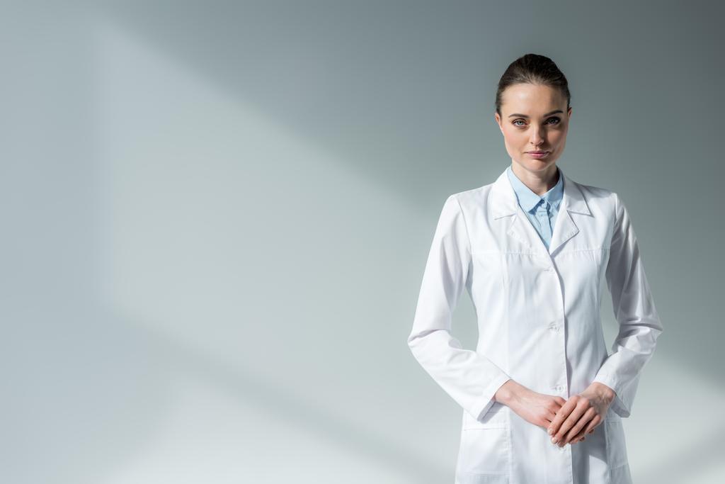 médico femenino serio en bata blanca mirando a la cámara en gris
 - Foto, imagen