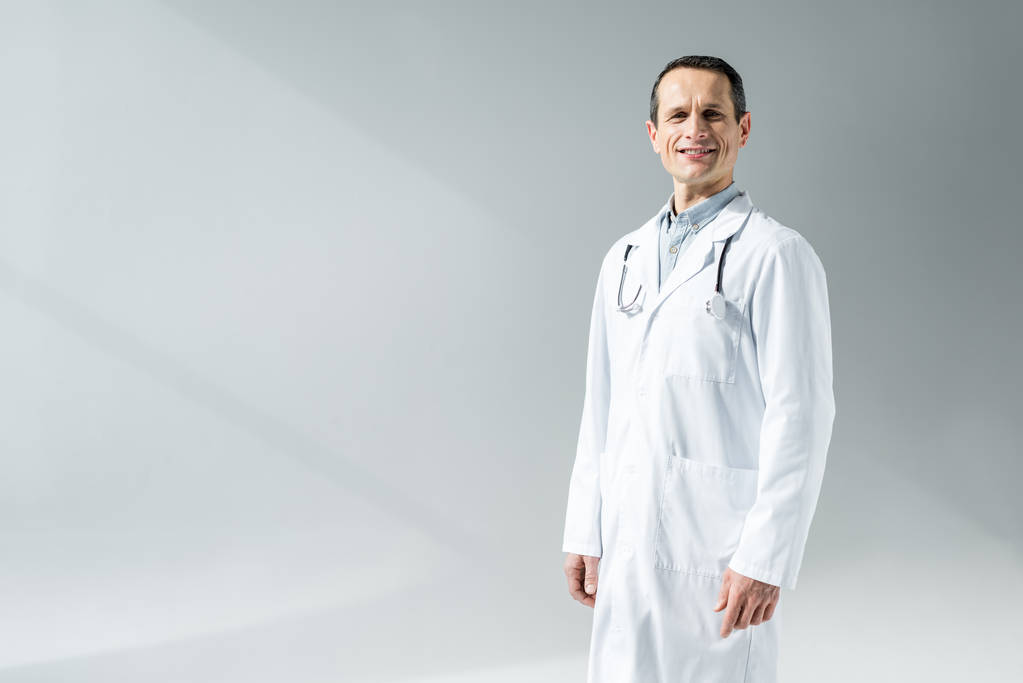 Lächelnder erwachsener Arzt im weißen Mantel, der in die Kamera schaut, isoliert auf grau - Foto, Bild