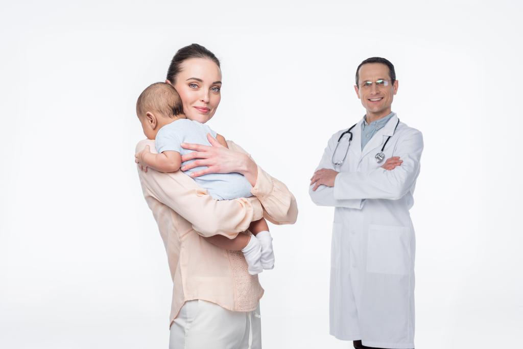 äiti syleilee lastaan, kun lastenlääkäri seisoo taustalla eristyksissä valkoisella
 - Valokuva, kuva