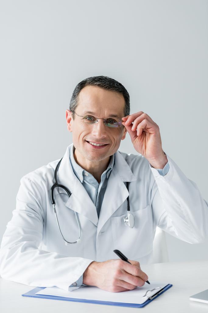 médico adulto feliz sentado en el lugar de trabajo y mirando a la cámara mientras escribe en portapapeles aislado en blanco
 - Foto, imagen