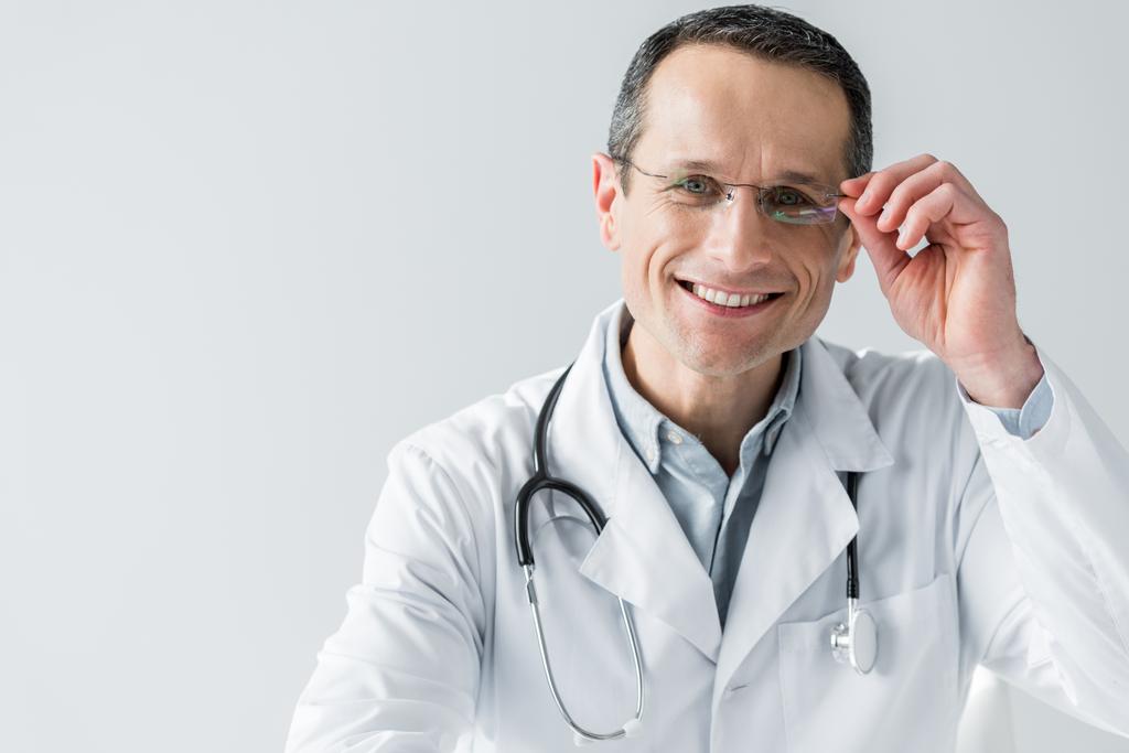 счастливый взрослый врач сидит на рабочем месте и смотрит на камеру изолированы на белом
 - Фото, изображение