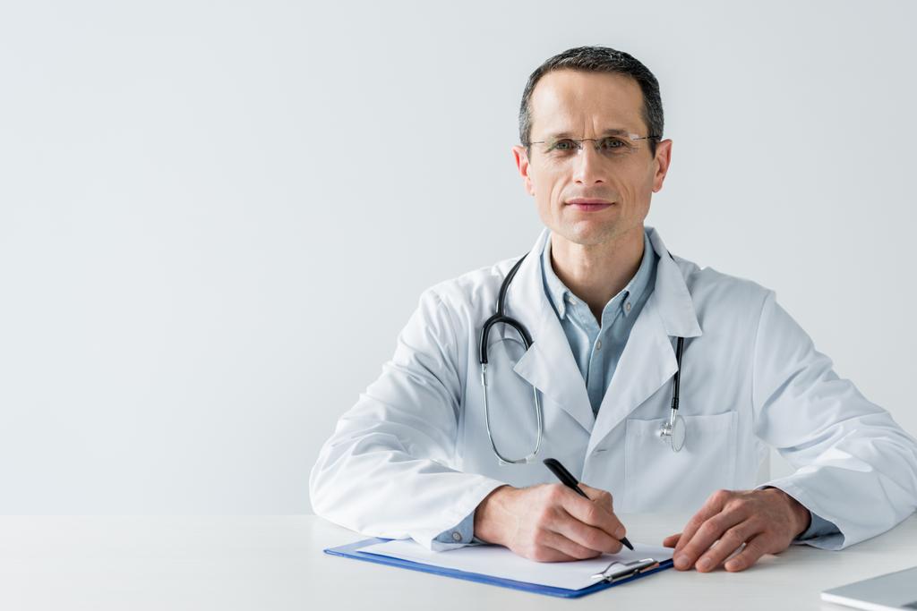 guapo médico adulto sentado en el lugar de trabajo y escribir diagnóstico en portapapeles aislado en blanco
 - Foto, imagen