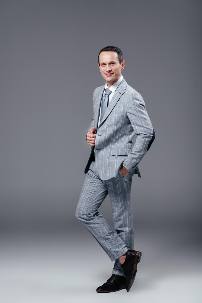 sonriente adulto hombre de negocios en traje elegante mirando a la cámara en gris
 - Foto, Imagen
