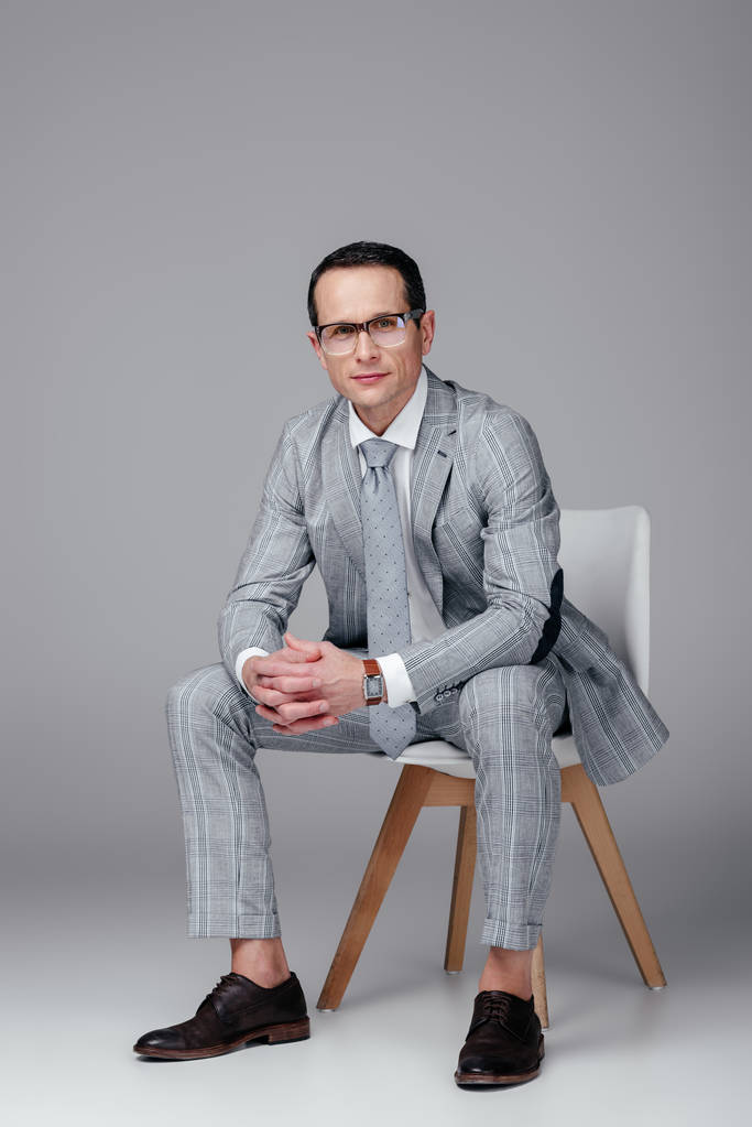 homem de negócios adulto sorridente em terno elegante sentado na cadeira e olhando para a câmera no cinza
 - Foto, Imagem