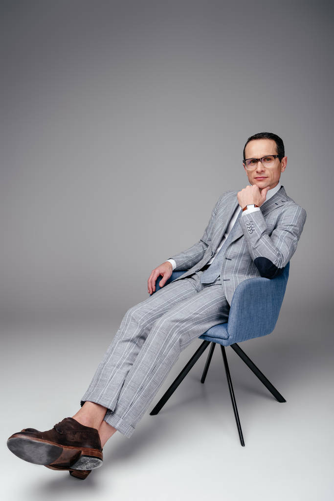 homem de negócios adulto em terno elegante sentado na cadeira com a mão no queixo no cinza
 - Foto, Imagem