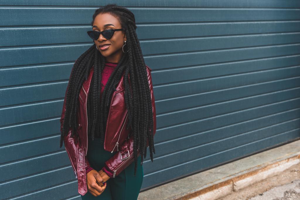 beautiful smiling young african american woman in fashionable jacket and sunglasses posing outside - Photo, Image