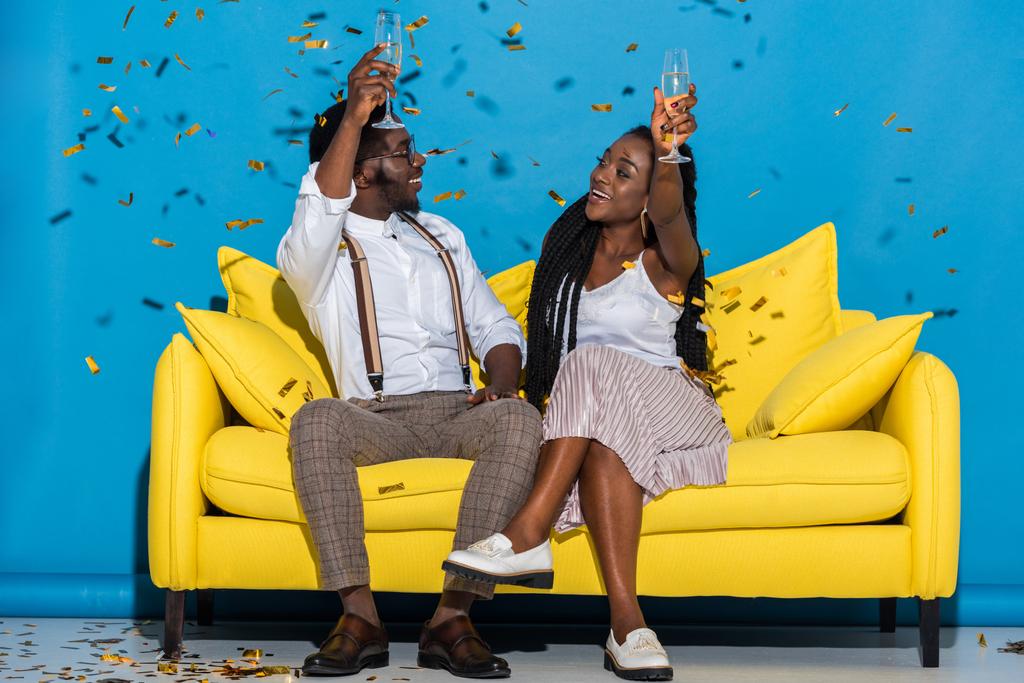 vrolijke stijlvolle jonge Afrikaanse Amerikaanse echtpaar holding glazen wijn en glimlachen elkaar zittend op de Bank onder dalende confetti - Foto, afbeelding
