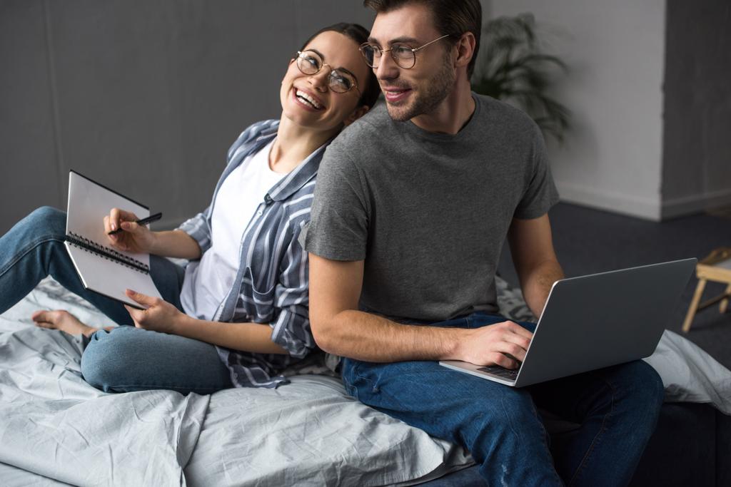 Счастливая пара в очках пишет в блокноте и работает на ноутбуке, сидя на кровати
 - Фото, изображение