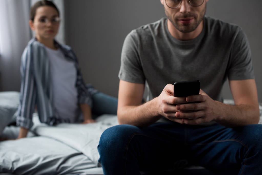 Nainen katselee miestä istuu sängyllä ja käyttää älypuhelinta
 - Valokuva, kuva