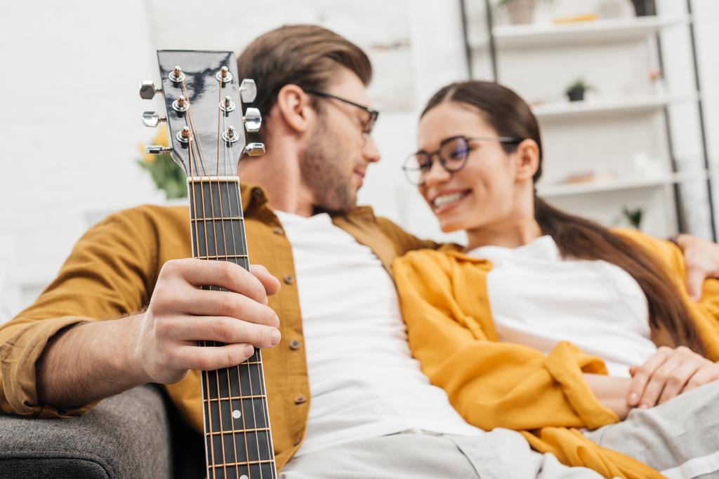 улыбающаяся пара с гитарой, сидящей дома на диване
 - Фото, изображение