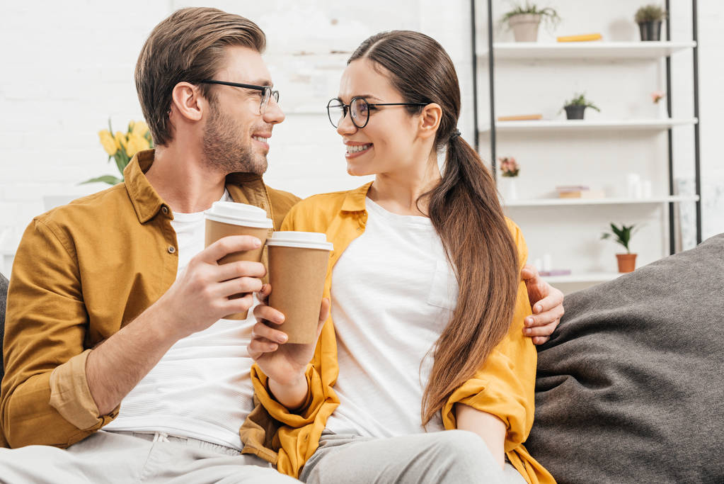 young happy couple flirting and clinking paper cups of coffee on couch - Photo, Image