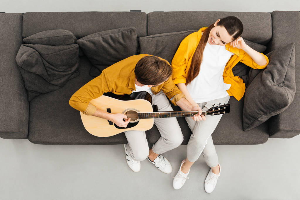 Вид сверху на человека, играющего на гитаре со своей подругой на диване дома
 - Фото, изображение