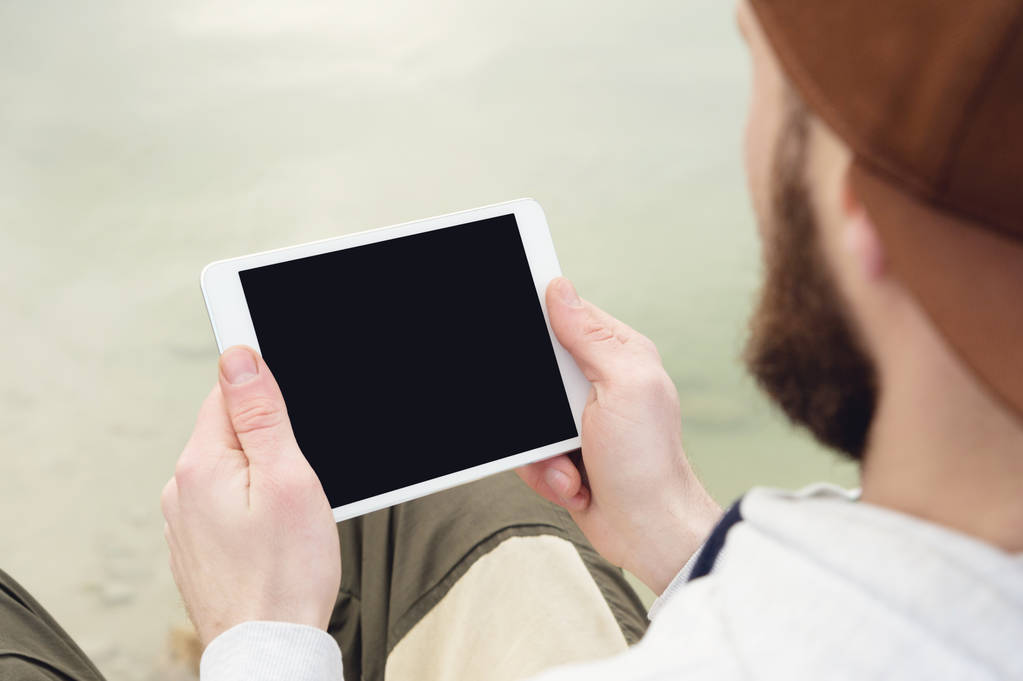 Primer plano de una horda en una gorra marrón al aire libre sostiene una tableta blanca PC en sus manos. Un hombre barbudo mira la tablilla. OTS vista desde detrás del hombro
 - Foto, Imagen